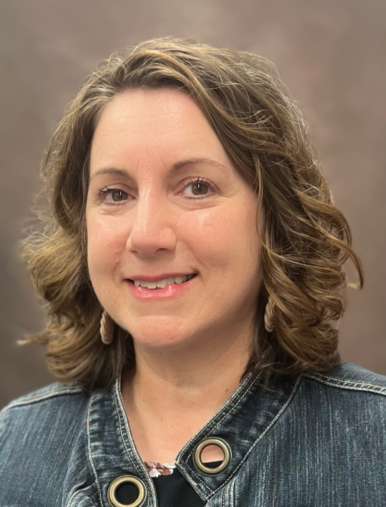 A woman with short curly hair, wearing a denim jacket and earrings, smiles at the camera against a plain background.